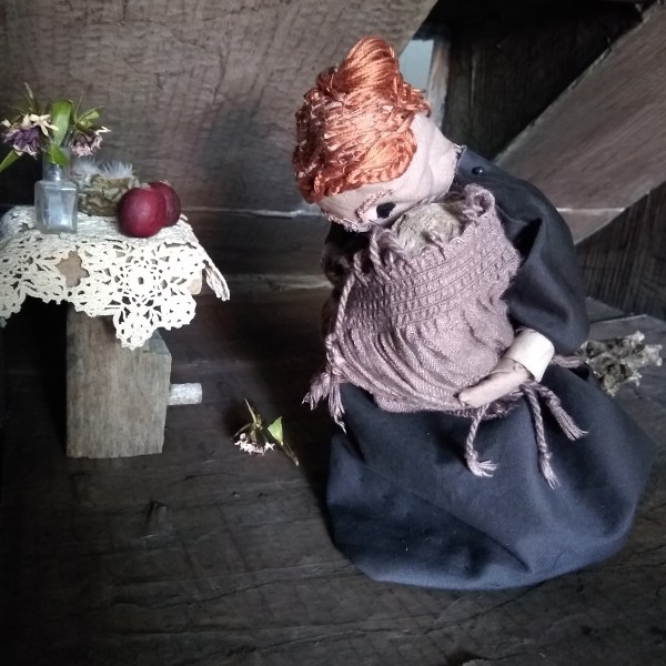A cloth puppet woman wears an old-fashioned grey dress as she kneels on a bare floor, she holds a baby wrapped in a shawl in her arms. She is in a dark attic room with a sloping roof. Beside her is a small table with a lace cover, a vase of flowers and two small red crab apples.