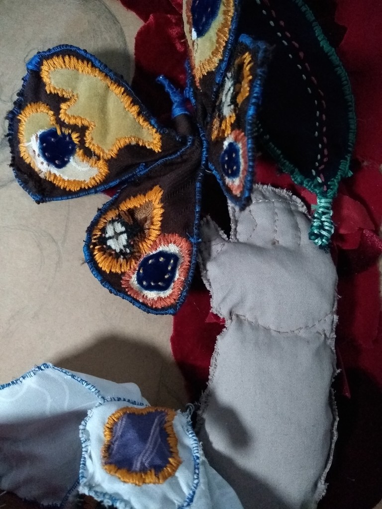 A child's hand made from cloth reaches towards a cloth butterfly, embroidered with blue, brown and gold spots. There are cloth flowers and leaves in the background, resting on brown paper and red velvet. Faint drawing marks can be seen on the paper.