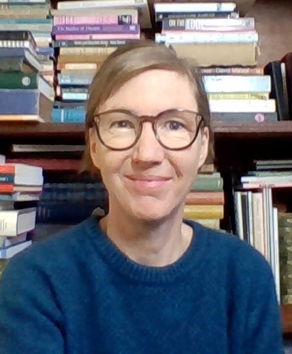 In this image, I am wearing reading glasses and a blue jumper, and smiling while sitting in front of a bookshelf.